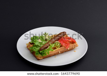 Classic sausage on waffles breakfast with tomatoes, salad and sauce served on a white plate on black background. Food photography concept.