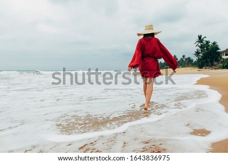 Picture of happy woman running along seashore. Rest or enjoying vacation. Activity on island in hot country. Young woman in motion. Wear red dress and barefeet