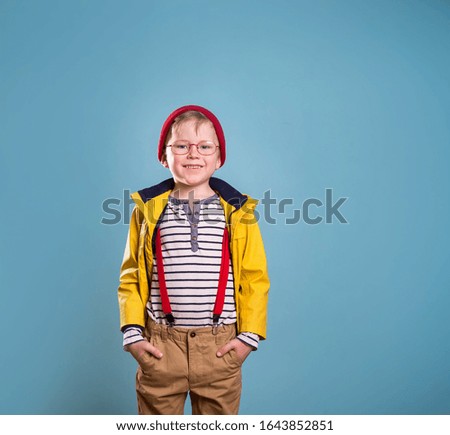 Stylish boy in striped shirt and yellow coat and red hat isolated on blue background. Confidant school kid with hands in pockets. 
