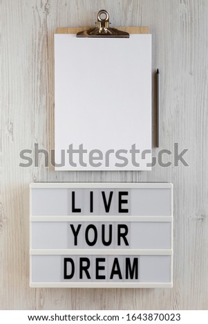 'Live your dream' words on a lightbox, clipboard with blank sheet of paper on a white wooden surface, top view. Overhead, from above, flat lay. 