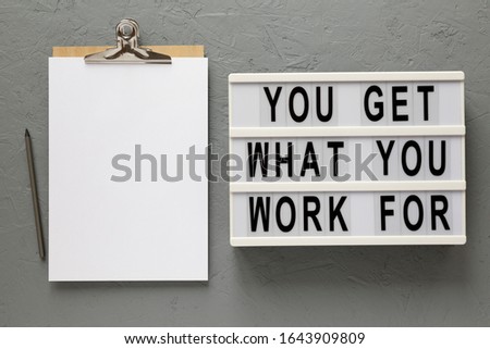 'You get what you work for' words on a lightbox, clipboard with blank sheet of paper on a gray surface, top view. Overhead, from above, flat lay. 