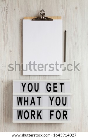 'You get what you work for' words on a lightbox, clipboard with blank sheet of paper on a white wooden background, overhead view. Top view, from above, flat lay. 