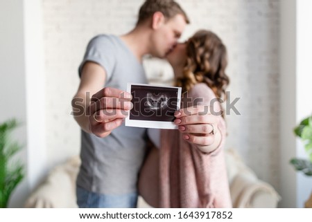 Future parents kissing each other in focus with their hands stretched out in front of them hold a picture of the child's ultrasound