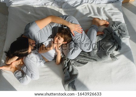 Young mother with her 4 years old little son dressed in pajamas are relaxing and playing in the bed at the weekend together, lazy morning, warm and cozy scene.