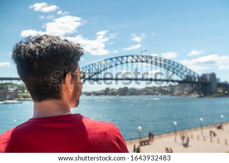 Man at Iconic Sydney Harbor Bridge over river water. Cityscape with buildings in CBD area. Tourist attraction in Australia. People at busy landmark. Black steel construction.