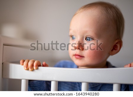 Beautiful blue eyed blonde baby boy looking to the left side and standing in the white cot. Baby is holding a cot rail and wearing blue outfit. Neutral face expression. Shallow depth of field portrait