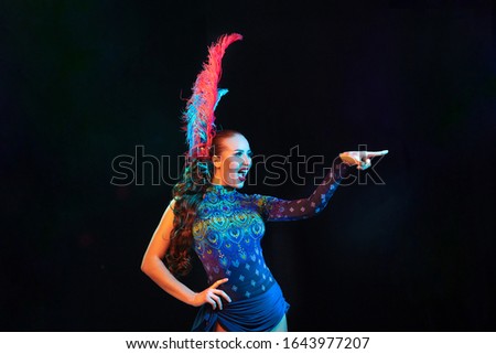 Pointing. Beautiful young woman in carnival, stylish masquerade costume with feathers on black background in neon light. Copyspace for ad. Holidays celebration, dancing, fashion. Festive time, party.
