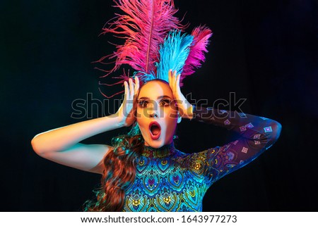 Astonished. Beautiful young woman in carnival, stylish masquerade costume with feathers on black background in neon light. Copyspace for ad. Holidays celebration, dancing, fashion. Festive time, party