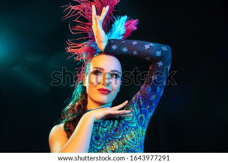 Flashlight. Beautiful young woman in carnival, stylish masquerade costume with feathers on black background in neon light. Copyspace for ad. Holidays celebration, dancing, fashion. Festive time, party