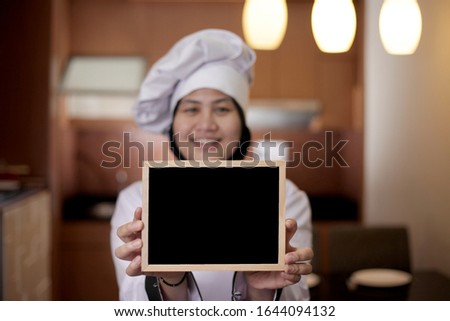 Portrait of Asian female chef looks happy and proud showing empty blackboard, presenting blank writing template, copy space meal menu concept