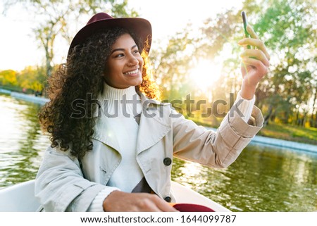 Cheerful young african woman wearing coat riding in a boat in the park, taking a selfie