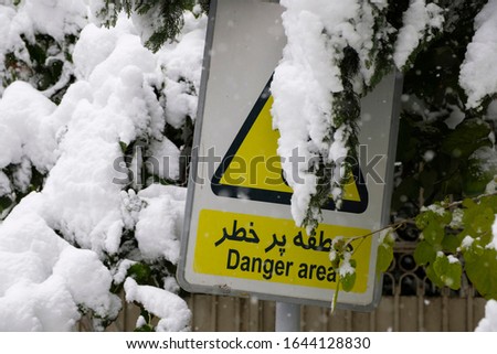 A traffic sign of caution and warning 'Danger Area' covered by heavy snow and a bent branch.