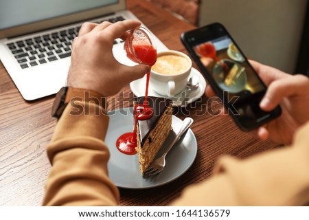 Male blogger taking photo of dessert and coffee at table in cafe, closeup Royalty-Free Stock Photo #1644136579