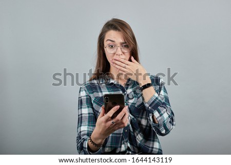 Young brunette girl blue green in checked shirt on grey background, shocked woman with glasses is talking on phone that looks like an iPhone