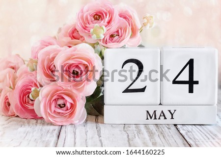 White wood calendar blocks with the date May 24th and pink ranunculus flowers over a wooden table. Selective focus with blurred background. 
