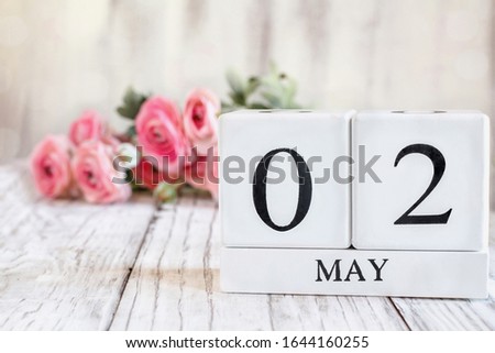 White wood calendar blocks with the date May 2nd. Selective focus with pink ranunculus in the background over a wooden table.