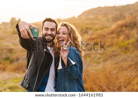 Beautiful young happy stylish couple wearing jackets standing at the beach, taking a selfie