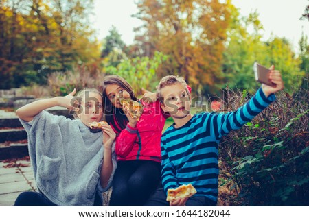 Group of a three children taking selfie by smart phone while eating pizza outdoors, having fun together. Kids shoving gesture thumb up.