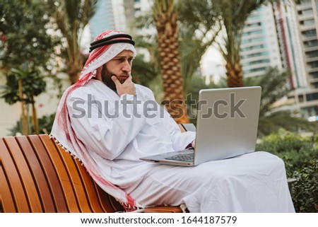 A man with a beard in Arabic folk costume is sitting on a bench and holding a laptop in his lap