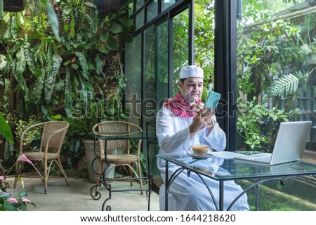 Arab muslim businessmen wear white traditional clothing in hand use smart phone and open laptop on coffee glass table