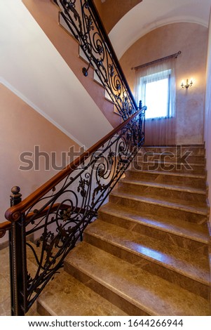 Cast-iron railings are forged in the form of a plant pattern. Handrail in lacquered mahogany. Marble steps with a pattern. Beige walls with a pinkish tinge