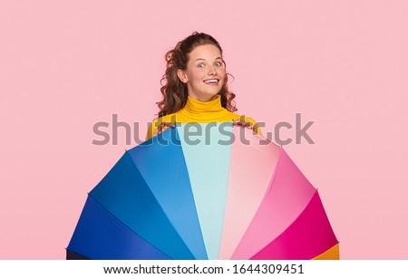 Delighted young woman peeking out from behind bright multicolored umbrella during summer vacation against pink background