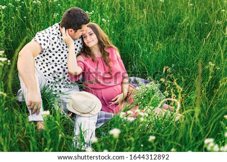 meadow with tall grass and the future parents sit on coverlet. bag with a bouquet of flowers and boater.