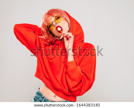 Young beautiful hipster bad girl in trendy red summer red hoodie and earring in her nose.Sexy carefree woman posing in studio on gray background in wig.Hot model licking round sugar candy Royalty-Free Stock Photo #1644383140