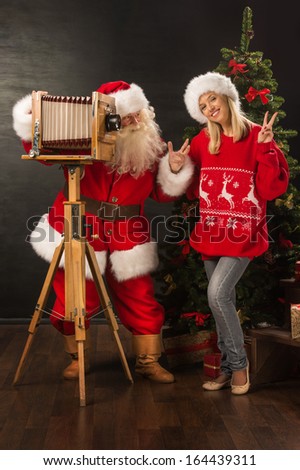 Santa Claus taking picture of cheerful woman with old wooden camera at home near Christmas tree