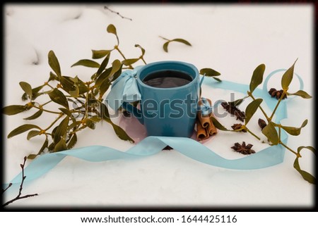 A blue cup with tea on a saucer stands on a white snow. Nearby scattered star anise, lies a blue ribbon and green leaves of mistletoe.