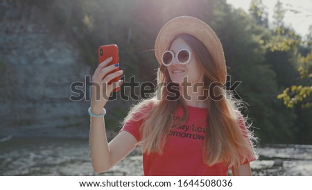 Smiling woman with hat wear sunglasses stand use smartphone near river forest tourism nature mountain cellphone summer enjoy mobile landscape vacation lake field happy outdoor outside slow motion