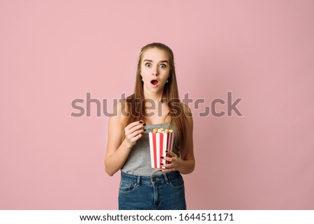 Photo of trendy scared blonde terrified girl eating pop corn wearing gray t-shirt watching movie while isolated with pink pastel background. Template for a blog about movies or movie theater ads