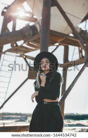 A long haired blonde woman, dressed all in black, wearing a black fedora type hat, black sun glasses and a knee high black dress, posing nonchalantly in front of a distressed industrial silo