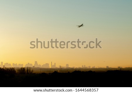 A plane is silhouetted against a colorful Yellow and Orange sunrise as it takes off from the Miami airport with the city in the background.