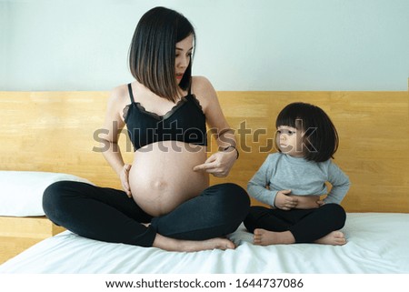 Mother and daughter sit on bed showing  tummy.