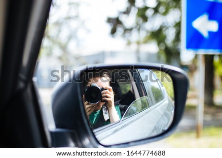 A woman is smiling and taking photos with a camera from car window looking through car mirror.   Copy space. Close up. Selective focus. Travel concept.