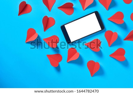 Black smartphone with isolated white screen for text, picture, photo and other graphics near scattered red paper hearts lies on blue table. Valentines day and love concept. Space for text. Top view