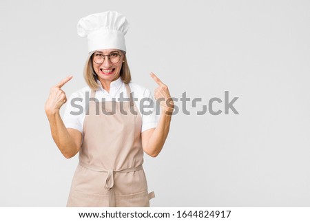 middle age baker woman with a bad attitude looking proud and aggressive, pointing upwards or making fun sign with hands against flat wall