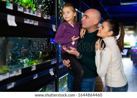 Happy family with cute preteen daughter choosing tropical fish for home aquarium in pet shop