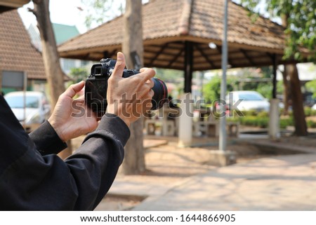 The person holding a large black camera standing in the garden. He is going to take a photo of the scenery around him.