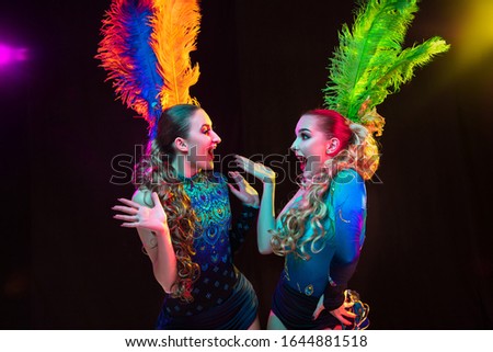 Surprised. Beautiful young women in carnival, stylish masquerade costume with feathers on black background in neon light. Copyspace for ad. Holidays celebration, dancing, fashion. Festive time, party.