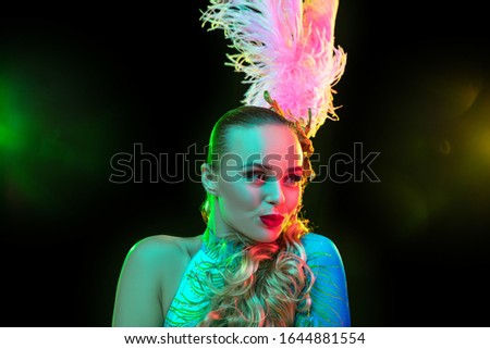 Cute. Beautiful young woman in carnival, stylish masquerade costume with feathers on black background in neon light. Copyspace for ad. Holidays celebration, dancing, fashion. Festive time, party.
