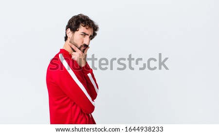 young bearded man back view looking serious, thoughtful and distrustful, with one arm crossed and hand on chin, weighting options against copy space wall