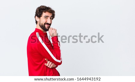young bearded man back view looking happy and smiling with hand on chin, wondering or asking a question, comparing options against copy space wall