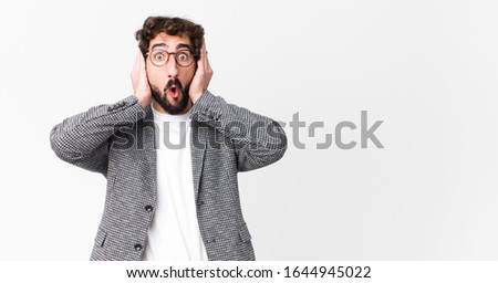 young crazy businessman looking unpleasantly shocked, scared or worried, mouth wide open and covering both ears with hands against flat wall