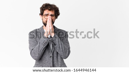 young crazy businessman feeling worried, hopeful and religious, praying faithfully with palms pressed, begging forgiveness against flat wall