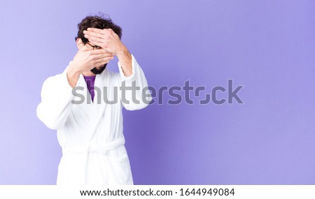 young bearded man wearing bathrobe covering face with both hands saying no to the camera! refusing pictures or forbidding photos