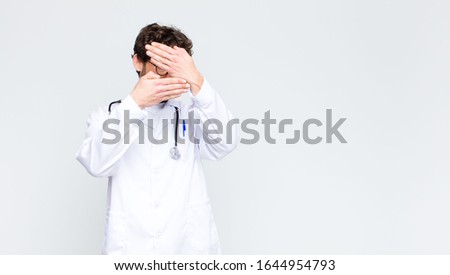 young doctor man covering face with both hands saying no to the camera! refusing pictures or forbidding photos against copy space wall