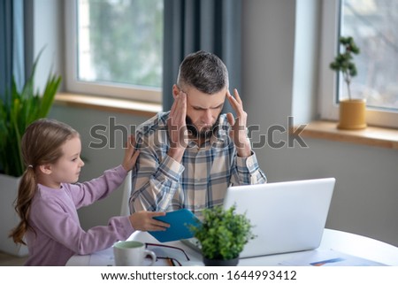 Fatigue. Tired dad sitting at a laptop with his eyes closed holding his hands to his head, next to a little daughter showing a tablet.