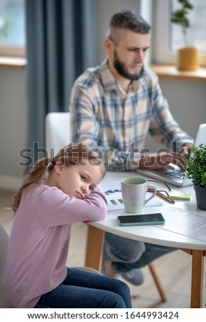 Lonely daughter. Sad daughter with her head resting on her hands and on the table, next to her concentrated concentrated dad working on a laptop.